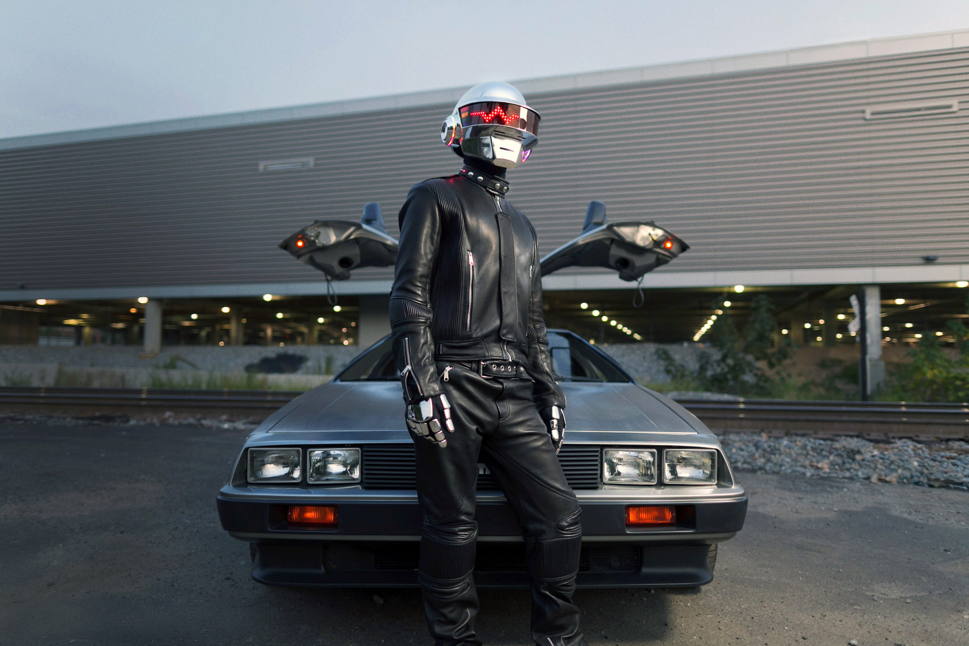 Helmeted robot figure leaning on a DeLorean at dusk
